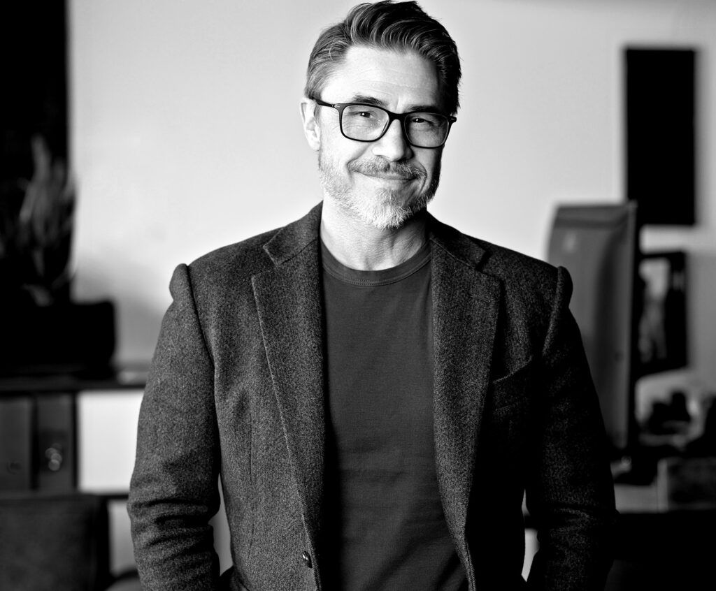 Black and white photo of a professional man with glasses. He is wearing a black t-shirt with a sports coat