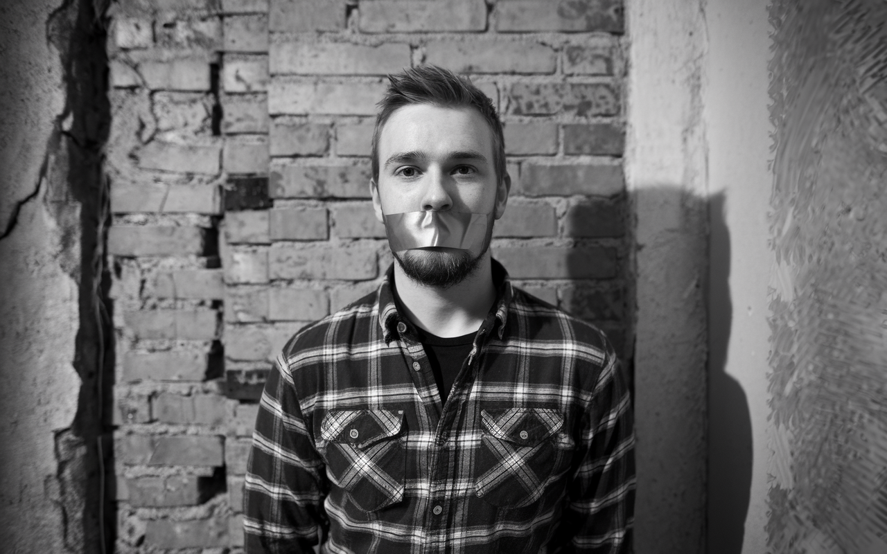Black and white photo of a guy with duct tape over his mouth standing up against a brick wall.
