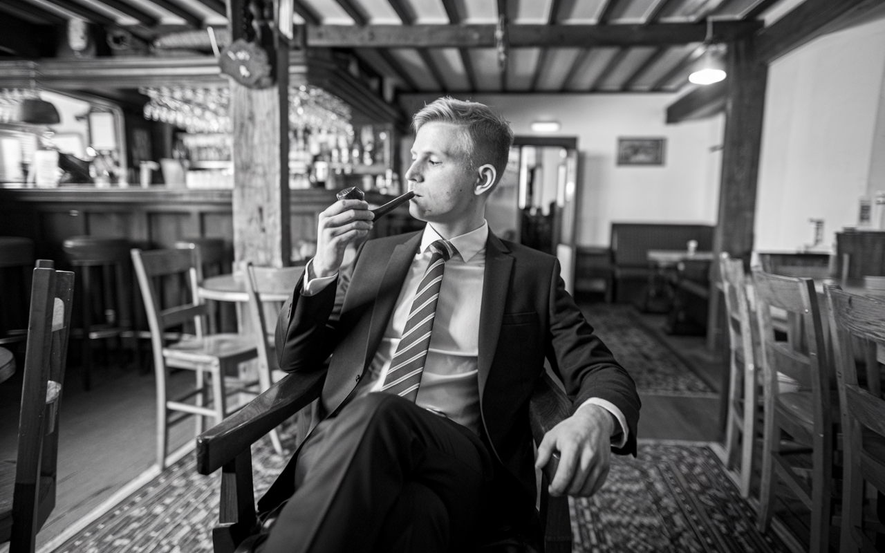 Black and white photo of a young man in a suite in a pub smoking a pipe in a suit and tie.
