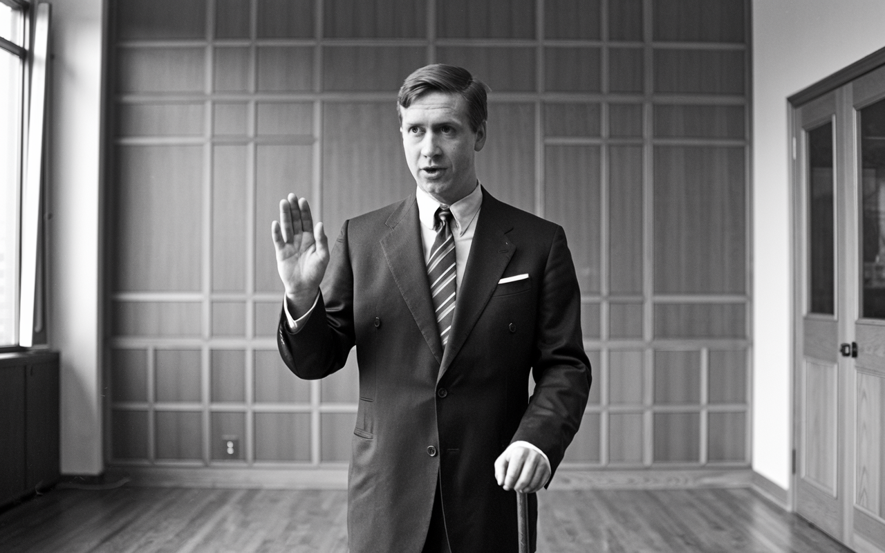 Black and white photo of a candidate with his right hand in the air standing in a room.