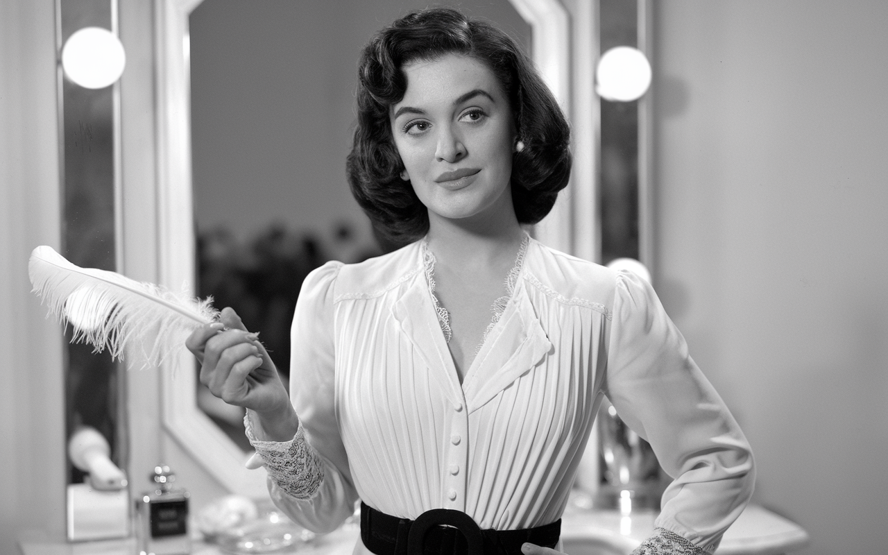 Black and white photo of a glamorous woman holding a feather pen looking off to the right.